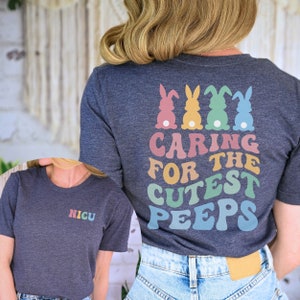 May include: A dark heather grey t-shirt with the text "NICU" on the front and "CARING FOR THE CUTEST PEEPS" on the back. The back design features four pastel colored bunnies.
