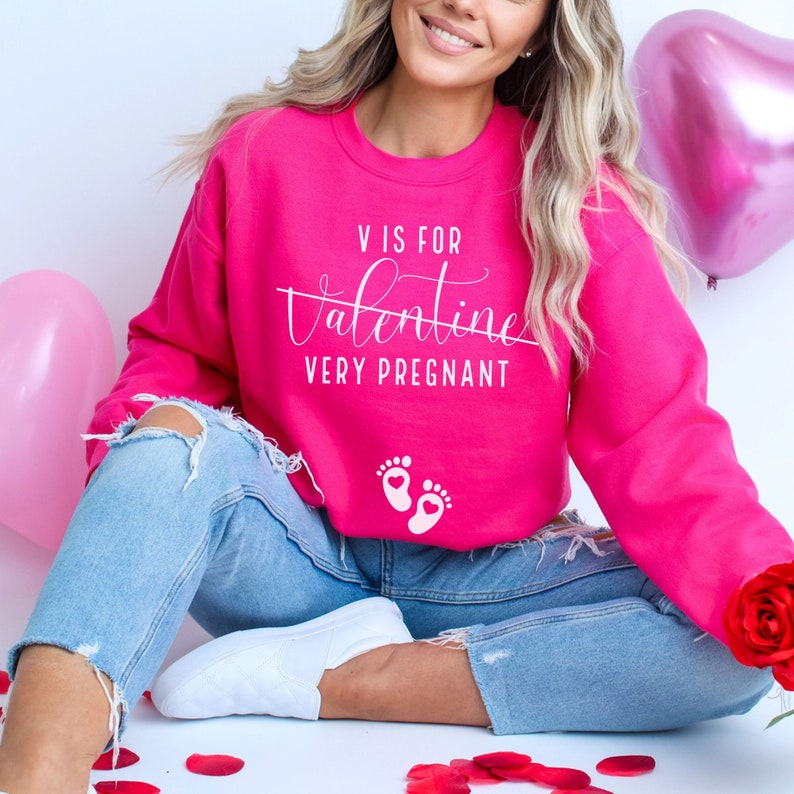May include: A pink sweatshirt with the text "V is for Valentine Very Pregnant" in white lettering. The sweatshirt also features a pair of white baby footprints with a heart in the center.