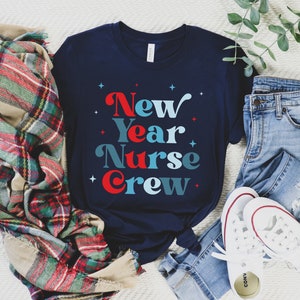 May include: A navy blue t-shirt with the text "New Year Nurse Crew" in red, blue, and white lettering. The shirt is laid out on a white surface with a plaid blanket, blue jeans, and white trainers.