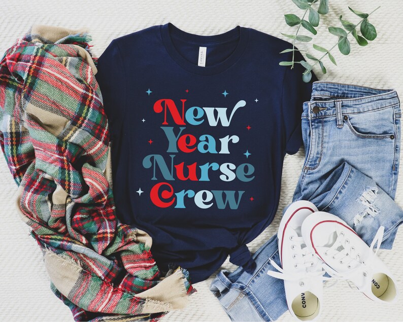 May include: A navy blue t-shirt with the text "New Year Nurse Crew" in red, blue, and white lettering. The shirt is laid out on a white surface with a plaid blanket, blue jeans, and white trainers.