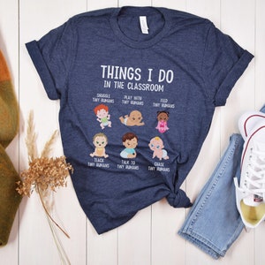 May include: A navy blue t-shirt with a white graphic that says "Things I do in the classroom" and features illustrations of babies with text describing activities like "Snuggle tiny humans", "Play with tiny humans", "Feed tiny humans", "Teach tiny humans", "Talk to tiny humans", and "Chase tiny humans".