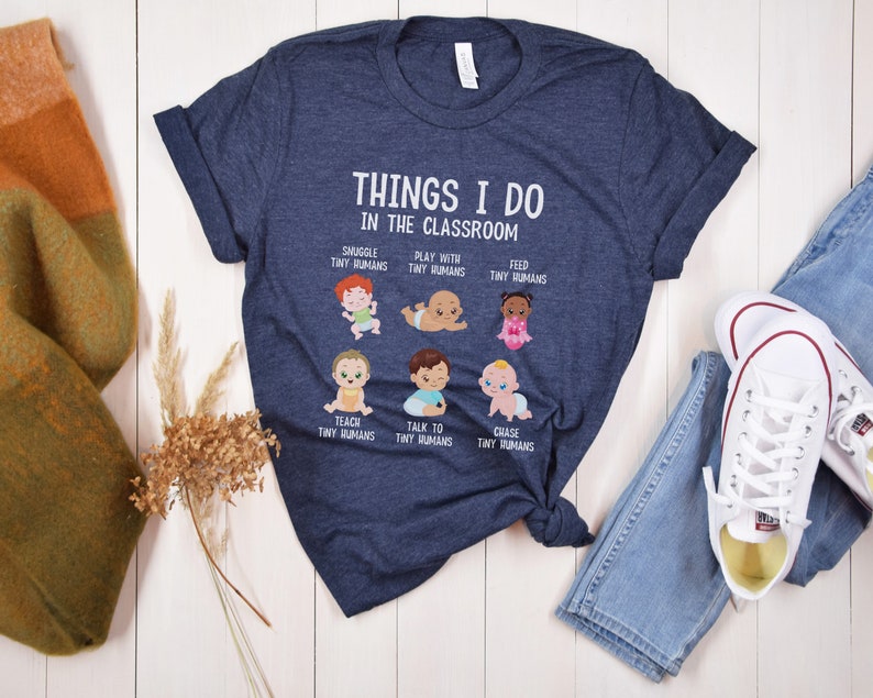 May include: A navy blue t-shirt with a white graphic that says "Things I do in the classroom" and features illustrations of babies with text describing activities like "Snuggle tiny humans", "Play with tiny humans", "Feed tiny humans", "Teach tiny humans", "Talk to tiny humans", and "Chase tiny humans".