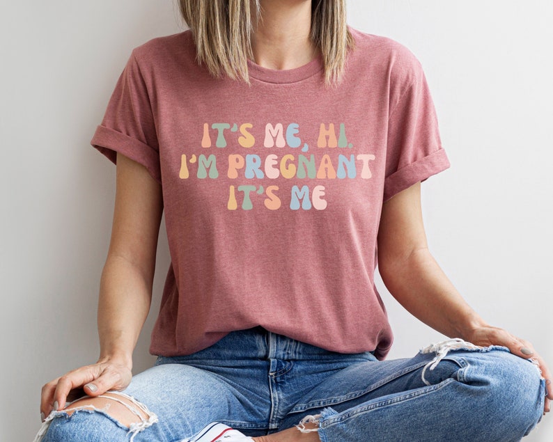 Puede incluir: Una mujer con una camiseta color marr&oacute;n rojizo con el texto "It's me, hi. I'm pregnant. It's me" en colores arco&iacute;ris.