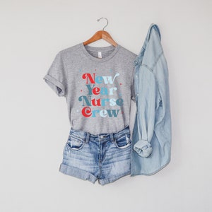 May include: A grey t-shirt with the text "New Year Nurse Crew" in blue, red and white lettering. The shirt is hanging on a wooden hanger and is paired with denim shorts and a light blue denim jacket.