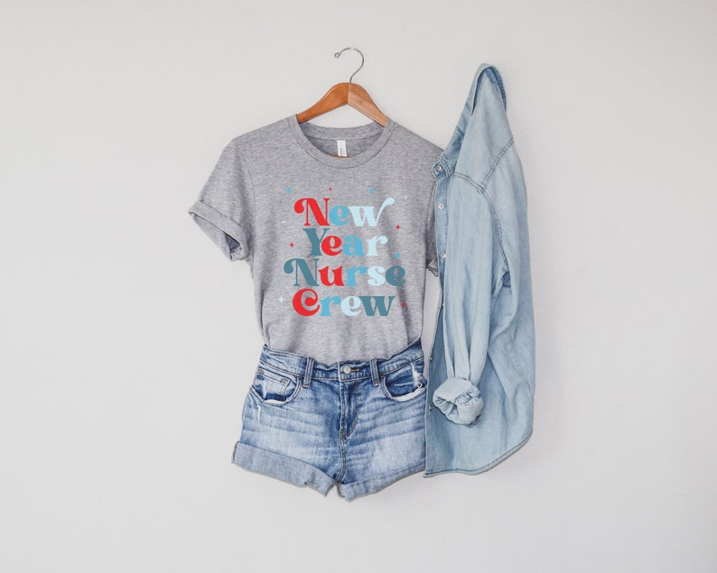 May include: A grey t-shirt with the text "New Year Nurse Crew" in blue, red and white lettering. The shirt is hanging on a wooden hanger and is paired with denim shorts and a light blue denim jacket.