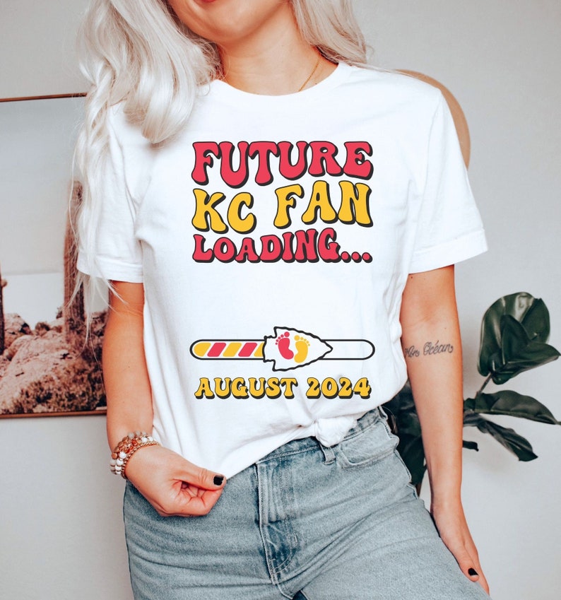 May include: White t-shirt with the text "FUTURE KC FAN LOADING..." in red and yellow letters. A progress bar with a red and yellow stripe is below the text, and the bar is filled with a red and yellow stripe. The progress bar ends with a red arrowhead with two yellow footprints inside. The text "AUGUST 2024" is below the progress bar.