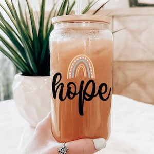 Puede incluir: Un tarro de vidrio transparente con tapa de bambú y una pajita. El tarro está lleno de un líquido naranja claro y tiene un arcoíris y la palabra "esperanza" impresa en la parte delantera.