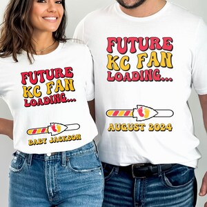 May include: White t-shirts with a red and yellow graphic that says "Future KC Fan Loading..." with a progress bar and a football-shaped arrow. The progress bar is filled with red and yellow stripes and has a red arrowhead with a yellow outline. The arrowhead has two yellow footprints. The text "August 2024" is below the progress bar on one shirt. The text "Baby Jackson" is below the progress bar on the other shirt.