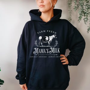 Op de afbeelding: Zwart hoodie met een witte afbeelding van een koe en de tekst "Farm Fresh Mama's Milk Locally Sourced - Always on Tap".