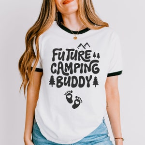 Może przedstawiać: Biała koszulka z czarnym wykończeniem wok&oacute;ł dekoltu. Koszulka ma czarny nadruk z napisem "Future Camping Buddy" z małym nadrukiem śladu stopy pod tekstem.