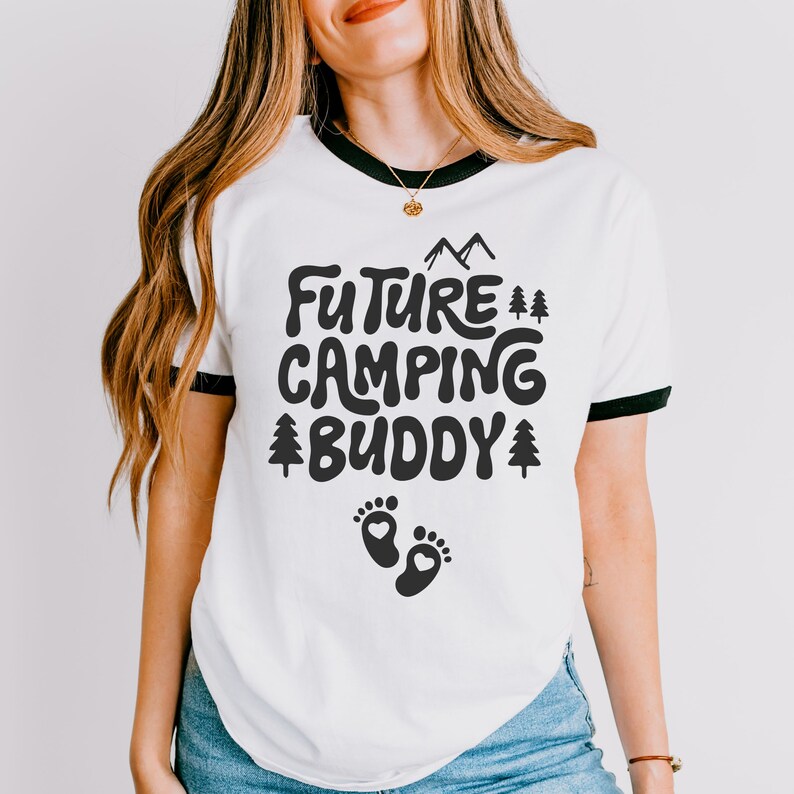 Może przedstawiać: Biała koszulka z czarnym wykończeniem wok&oacute;ł dekoltu. Koszulka ma czarny nadruk z napisem "Future Camping Buddy" z małym nadrukiem śladu stopy pod tekstem.