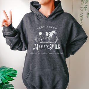 Op de afbeelding: Een donkergrijs hoodie met een wit grafisch ontwerp met de tekst "Farm Fresh Mama's Milk" met een koe-illustratie en de tekst "Locally Sourced - Always on Tap".