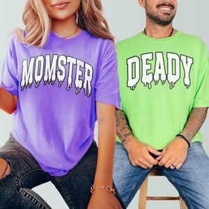 May include: Two people wearing matching Halloween shirts. The person on the left is wearing a purple shirt with the text "MOMSTER" in white dripping letters. The person on the right is wearing a green shirt with the text "DEADLY" in white dripping letters.