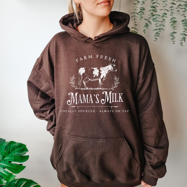 Op de afbeelding: Bruin hoodie met een wit grafisch ontwerp met de tekst "Farm Fresh Mama's Milk" met een koe-illustratie en de tekst "Locally Sourced. Always on Tap."