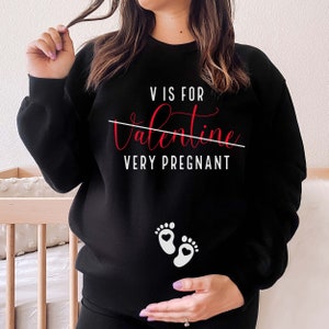 May include: Black sweatshirt with the text "V is for Valentine Very Pregnant" with a line through "Valentine" and two white baby footprints with red hearts on the bottom.