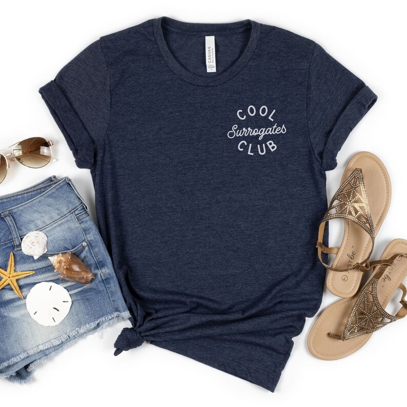 May include: Navy blue t-shirt with the text "Cool Surrogates Club" in a white, curved font. The shirt is paired with denim shorts, sunglasses, sandals, and seashells, suggesting a casual, beachy theme.