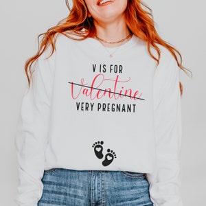 May include: White long-sleeve shirt with the text "V is for Valentine" with a line through "Valentine" and the text "Very Pregnant" below.  Two black footprints with a small heart in each are below the text.