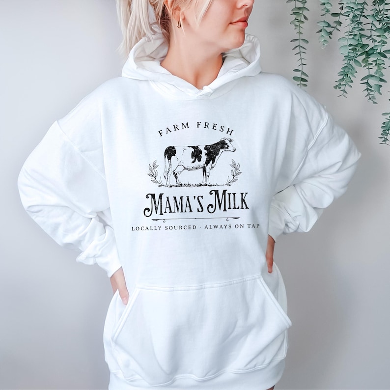 Op de afbeelding: Witte hoodie met een zwart-witte koe-illustratie en de tekst "Farm Fresh Mama's Milk Locally Sourced - Always on Tap".