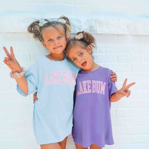 May include: Two young girls wearing matching light blue and purple t-shirts with the text "Lake Bum" in pink letters. The girls are smiling and making peace signs with their hands.