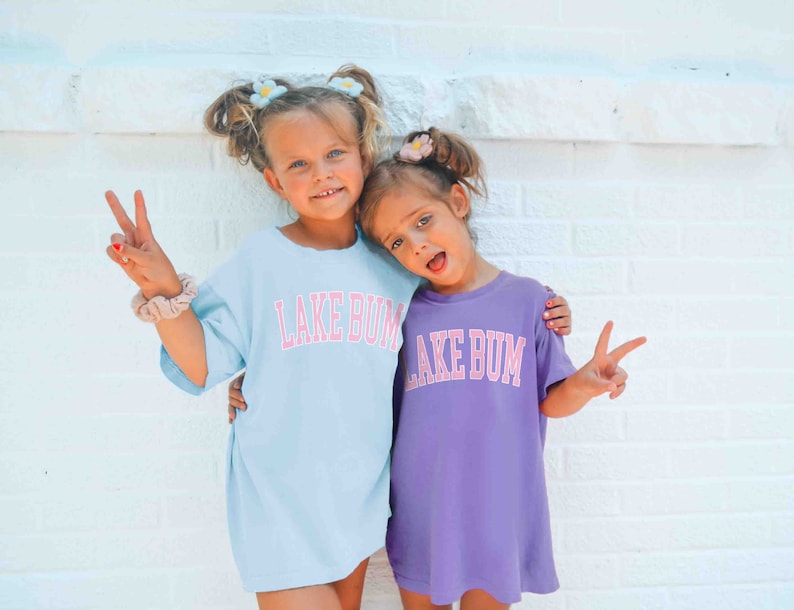 May include: Two young girls wearing matching light blue and purple t-shirts with the text "Lake Bum" in pink letters. The girls are smiling and making peace signs with their hands.