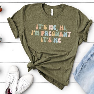 Puede incluir: Una camiseta verde con el texto "It's me, hi. I'm pregnant. It's me" en letras redondas y de colores.