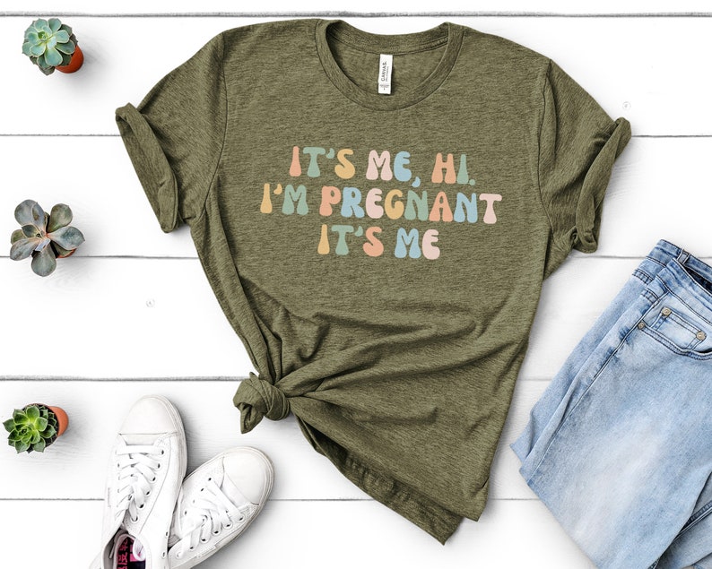 Puede incluir: Una camiseta verde con el texto "It's me, hi. I'm pregnant. It's me" en letras redondas y de colores.