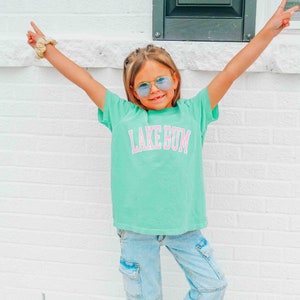 May include: A young person wearing a turquoise t-shirt with the text "LAKE BUM" in pink letters. They are wearing blue jeans and blue tinted sunglasses. The person is standing in front of a white brick wall.