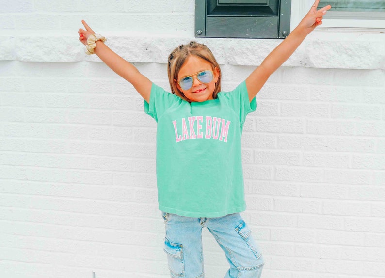 May include: A young person wearing a turquoise t-shirt with the text "LAKE BUM" in pink letters. They are wearing blue jeans and blue tinted sunglasses. The person is standing in front of a white brick wall.