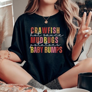 Puede incluir: Una camiseta negra con un dise&ntilde;o gr&aacute;fico colorido que dice "Crawfish, Hot Sauce, Mudbugs, Potatoes, Baby Bumps".