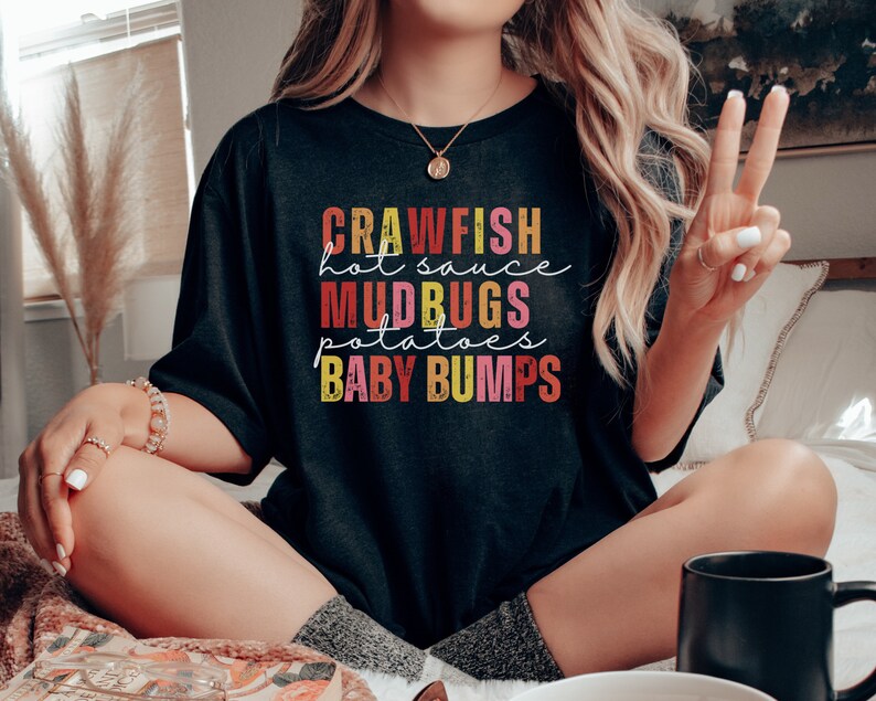 Puede incluir: Una camiseta negra con un dise&ntilde;o gr&aacute;fico colorido que dice "Crawfish, Hot Sauce, Mudbugs, Potatoes, Baby Bumps".