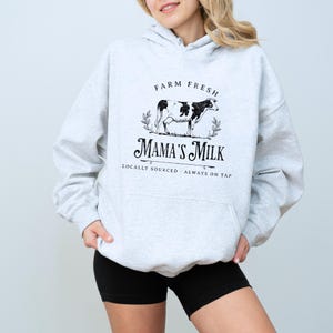 Op de afbeelding: Grijs hoodie met een zwarte afbeelding van een koe en de tekst "Farm Fresh Mama's Milk Locally Sourced - Always on Tap".