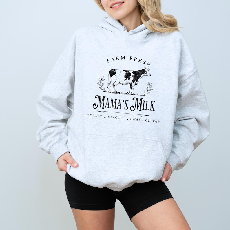 Op de afbeelding: Grijs hoodie met een zwarte afbeelding van een koe en de tekst "Farm Fresh Mama's Milk Locally Sourced - Always on Tap".