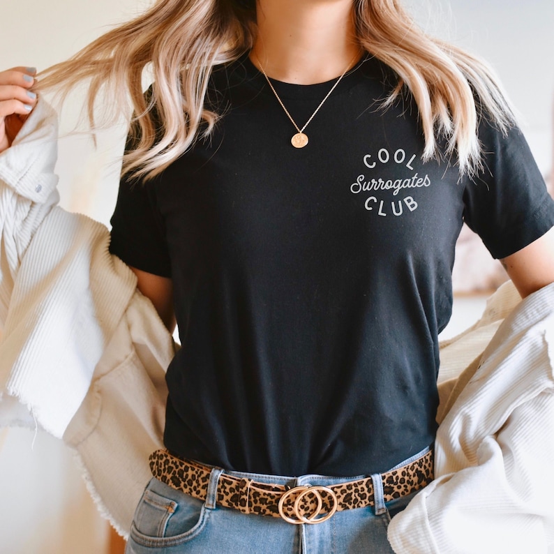 May include: Black t-shirt with the text "COOL Surrogates CLUB" in white script. The shirt is paired with a leopard print belt with gold double-circle buckle and blue jeans. A gold necklace is also visible.