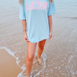 May include: A light blue t-shirt with the text "LAKE BUM" in pink lettering. The shirt is oversized and worn by a person standing on a sandy beach.