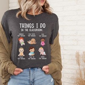 May include: A dark gray t-shirt with the text "THINGS I DO IN THE CLASSROOM" and illustrations of babies with text describing activities like "SNUGGLE TINY HUMANS", "PLAY WITH TINY HUMANS", "FEED TINY HUMANS", "TEACH TINY HUMANS", "TALK TO TINY HUMANS", and "CHASE TINY HUMANS".