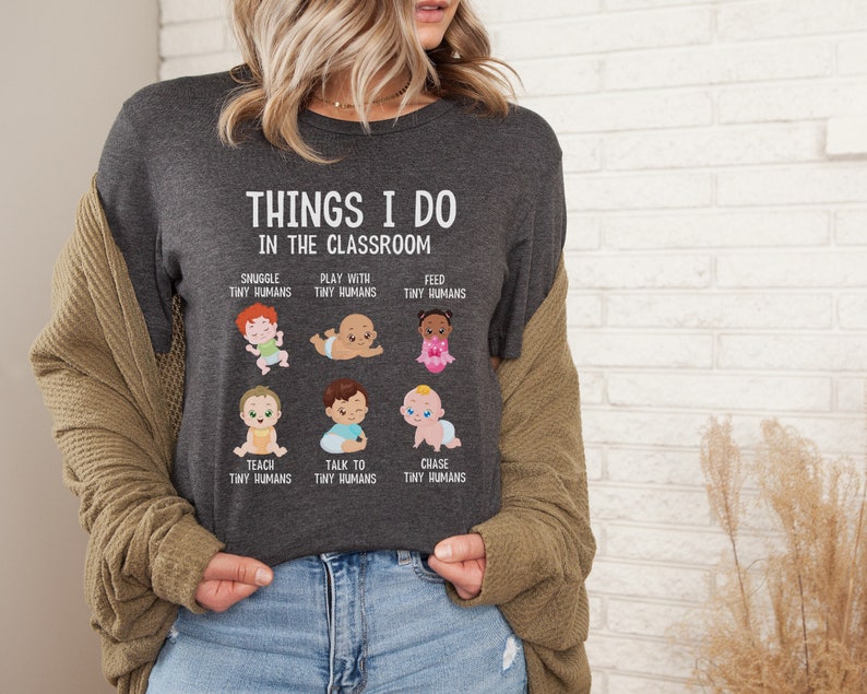 May include: A dark gray t-shirt with the text "THINGS I DO IN THE CLASSROOM" and illustrations of babies with text describing activities like "SNUGGLE TINY HUMANS", "PLAY WITH TINY HUMANS", "FEED TINY HUMANS", "TEACH TINY HUMANS", "TALK TO TINY HUMANS", and "CHASE TINY HUMANS".