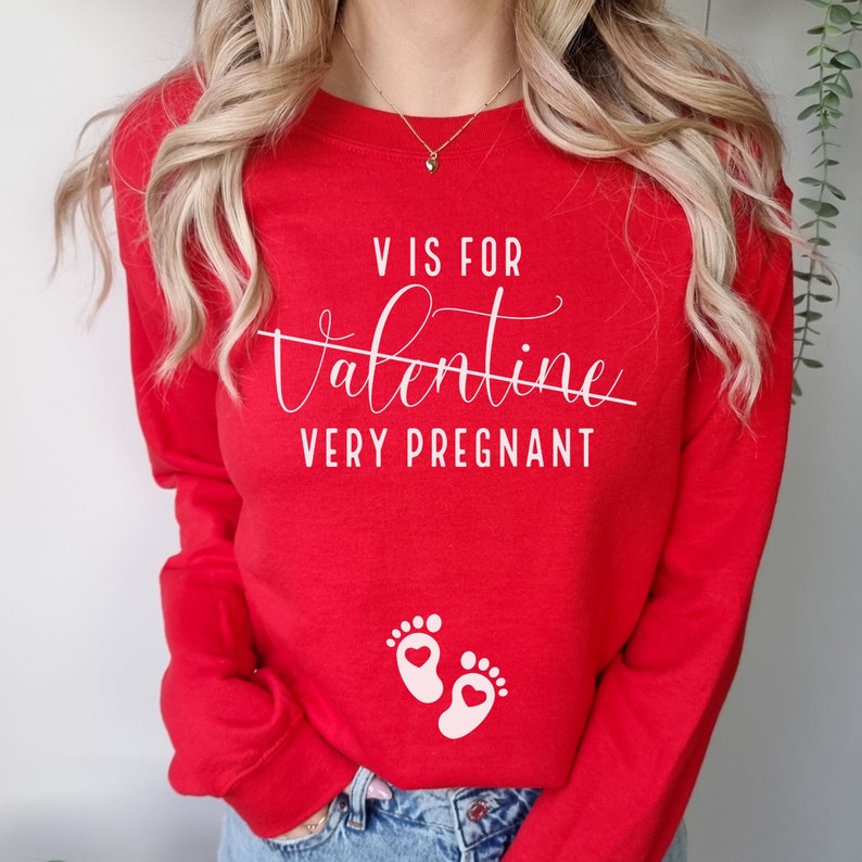 May include: Red long-sleeved sweatshirt with white text that reads "V is for Valentine Very Pregnant" and a pair of pink baby footprints with hearts.
