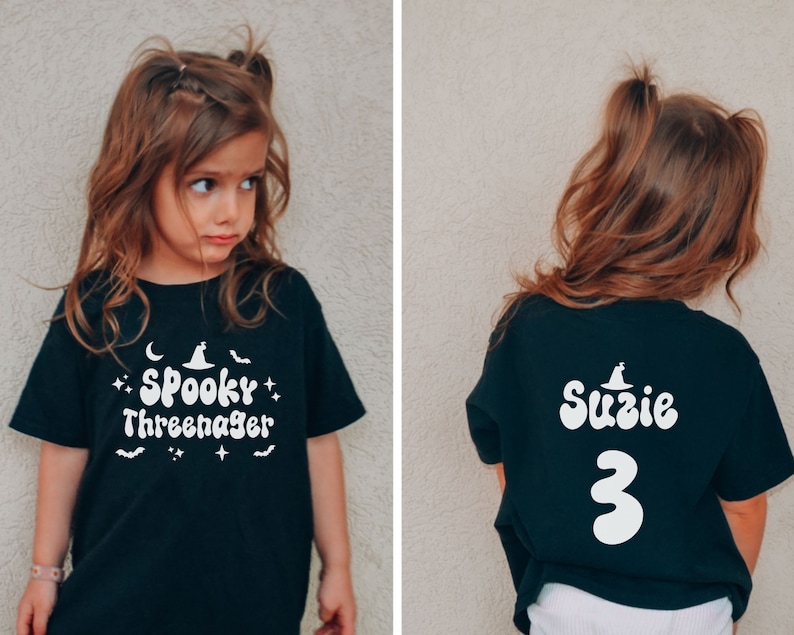 Op de afbeelding: Zwart T-shirt met witte tekst die "Spooky Three-nager" op de voorkant en "Suzie 3" op de achterkant zegt. Het shirt heeft een heksenhoed-afbeelding.