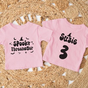 Op de afbeelding: Twee roze T-shirts met zwarte Halloween-ontwerpen. E&eacute;n shirt zegt "Spooky Three-nager" met een heksenhoed en sterren. Het andere shirt zegt "Suzie" met een heksenhoed en het nummer "3".