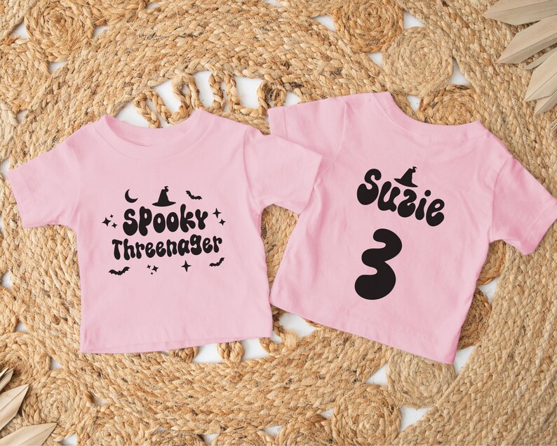 Op de afbeelding: Twee roze T-shirts met zwarte Halloween-ontwerpen. E&eacute;n shirt zegt "Spooky Three-nager" met een heksenhoed en sterren. Het andere shirt zegt "Suzie" met een heksenhoed en het nummer "3".