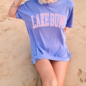 May include: A woman wearing a light purple t-shirt with the text "LAKE BUM" in pink letters. She is sitting on a sandy beach.
