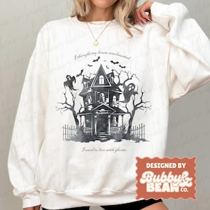 Pode incluir: Camisola branca com um gráfico de casa assombrada em preto e cinza, morcegos e fantasmas. O texto diz "I thought my house was haunted" e "I used to live with ghosts." Desenhado por Bubby & Bean Co.