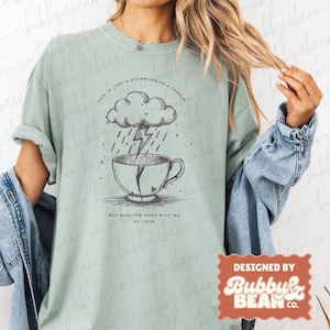 Pode incluir: Camiseta verde claro com um desenho de xícara de chá com uma nuvem de tempestade e um raio. O texto diz "THIS IS JUST A STORM INSIDE A TEACUP" e "BUT SHELTER HERE WITH ME, MY LOVE". A camiseta é rotulada como "comfort colors - bay".