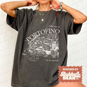 May include: Dark gray t-shirt with a white graphic of the town of Portofino, Italy, and the text "That view of PORTOFINO was on my mind." The shirt also has the text "Designed by Bubby & Bean Co."