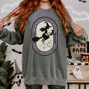 May include: Dark grey sweatshirt featuring a Halloween-themed graphic. The design includes a witch on a broom with a cat, and the text "They're burning all the witches, even if you aren't one, so light up". The sweatshirt has a crew neck and long sleeves.