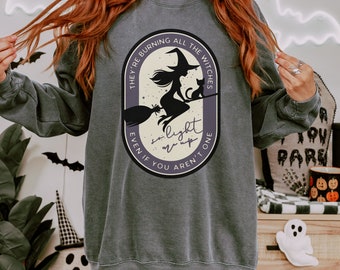 May include: Dark grey sweatshirt featuring a Halloween-themed graphic. The design includes a witch on a broom with a cat, and the text "They're burning all the witches, even if you aren't one, so light up". The sweatshirt has a crew neck and long sleeves.