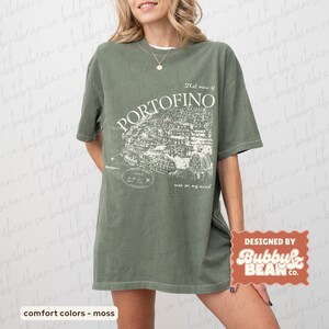 May include: Olive green t-shirt with a white graphic of Portofino, Italy, and the text "That view of Portofino was on my mind." The shirt also features the text "comfort colors - moss" and "DESIGNED BY Bubby & Bean Co."