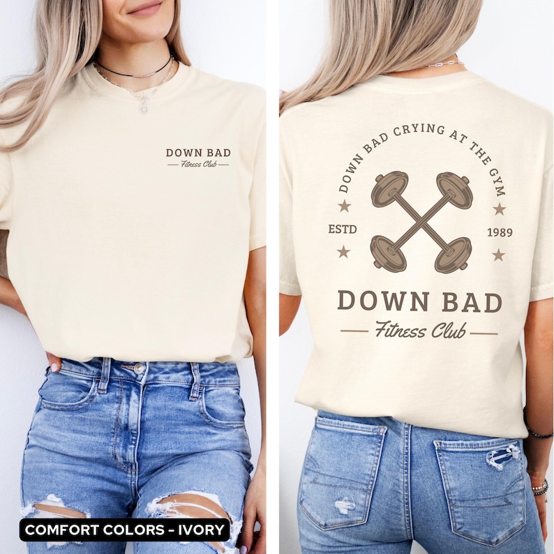 May include: Ivory t-shirt with a graphic of two crossed dumbbells and the text "Down Bad Fitness Club" on the back. The front of the shirt has the text "Down Bad Fitness Club" printed on it.