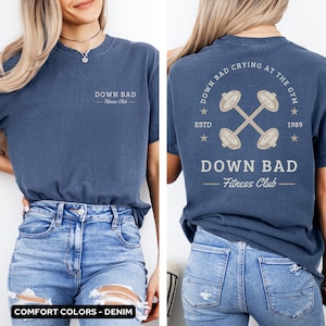 May include: A blue t-shirt with a white graphic design on the front and back. The front design says "Down Bad - Fitness Club". The back design says "Down Bad - Fitness Club" with a crossed dumbbell graphic and the text "Down Bad Crying At The Gym - Estd 1989".