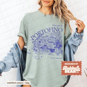 May include: A light green t-shirt with a blue graphic of the Italian town of Portofino. The text "That view of Portofino was on my mind" is printed above the illustration. The shirt is designed by Bubby & Bean Co.